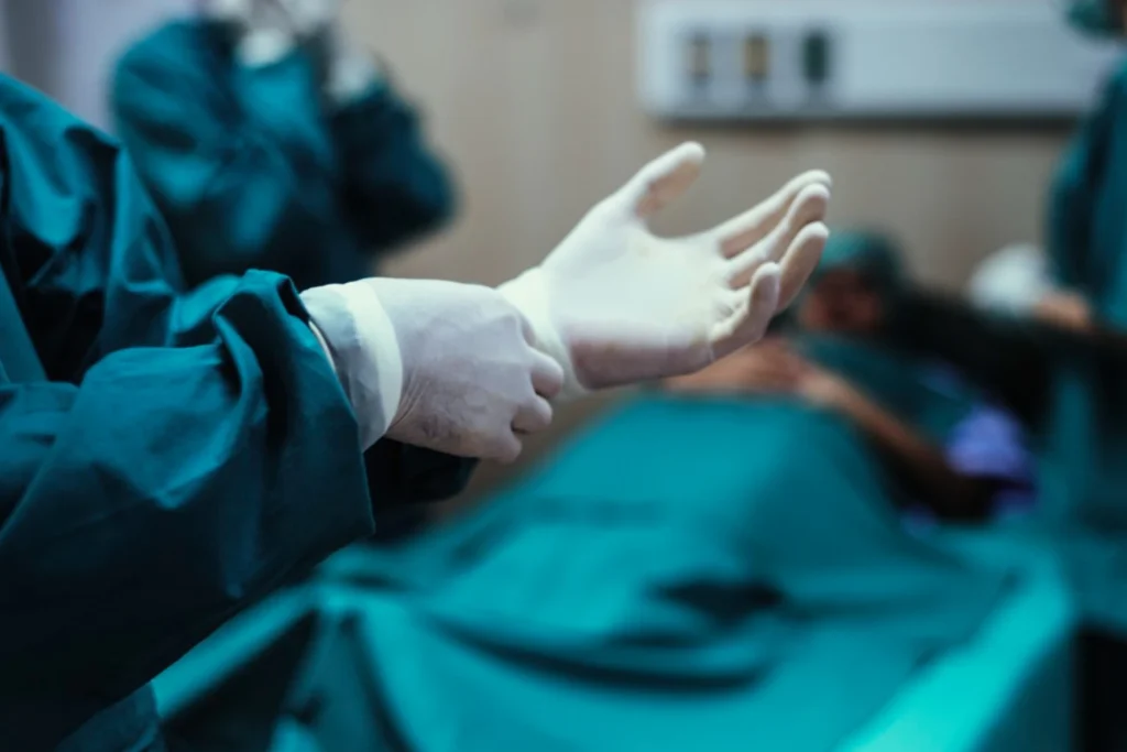Imagem de uma pessoa usando luvas e roupas cirúrgicas verdes em uma sala de cirurgia ilustrando como é feita a vasectomia.