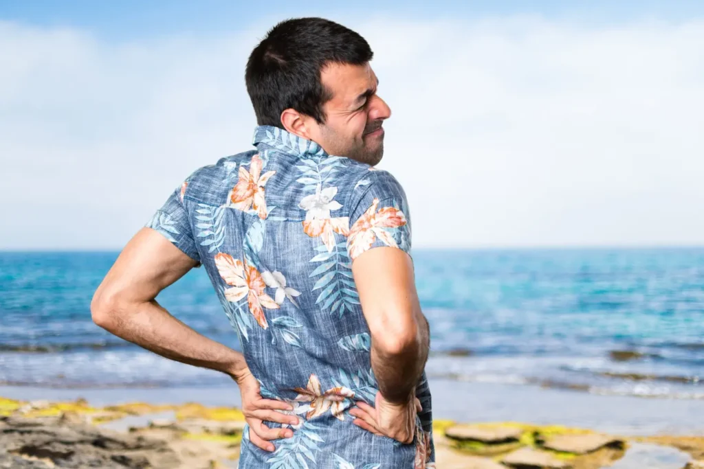 Imagem de homem branco de camisa florida na praia com as mãos na lombar ilustrando riscos de cálculos renal no verão.
